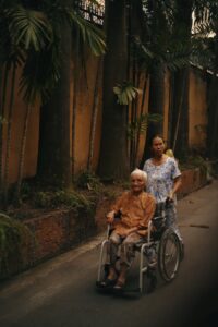 A caregiver pushing a senior woman in a wheelchair along a tropical street.